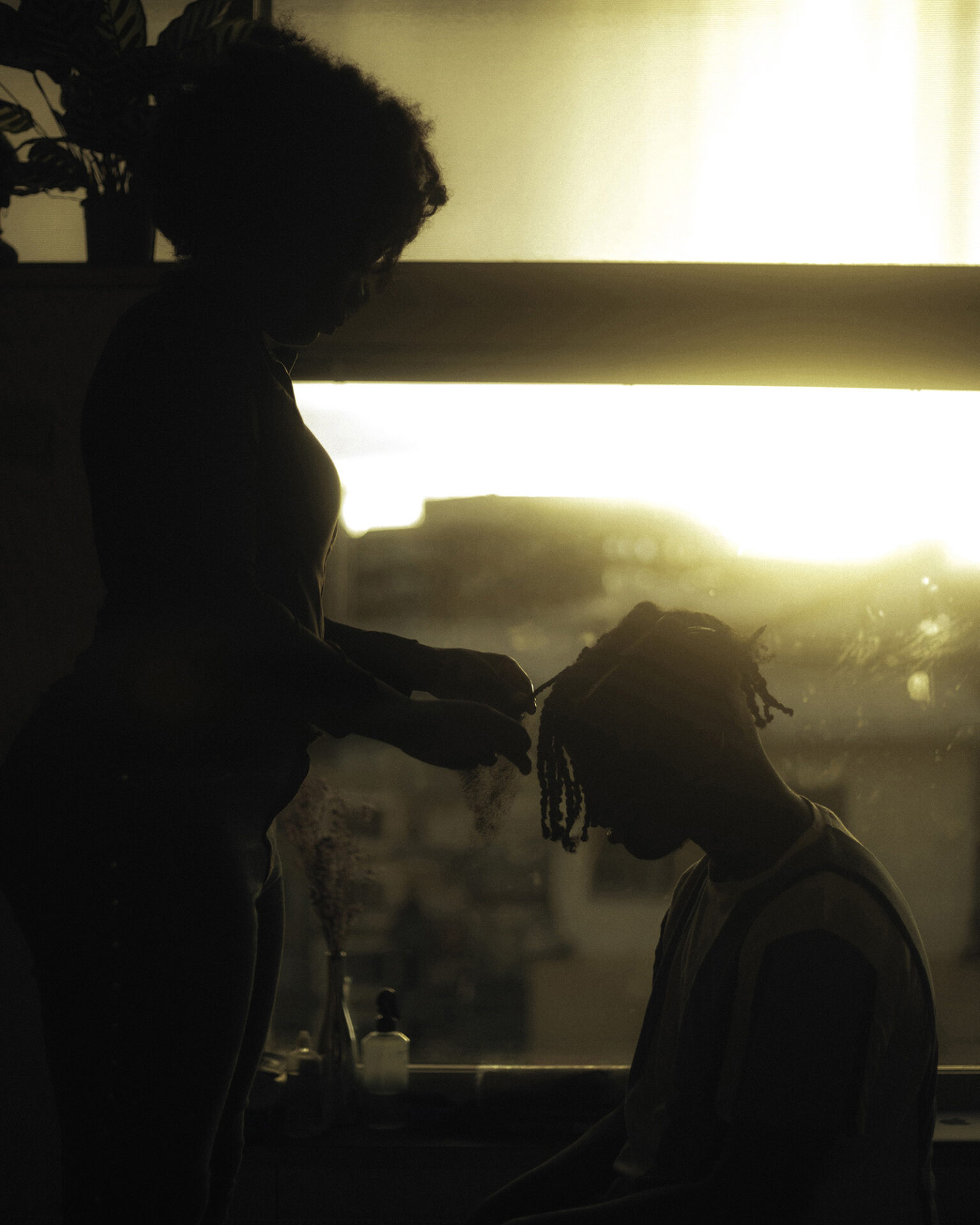 Photographer Ikram Abdulkadir's image features two people in silhouette in front of a window with sunshine pouring through it. One is standing, plaiting the hair of the second person, who is kneeling before them