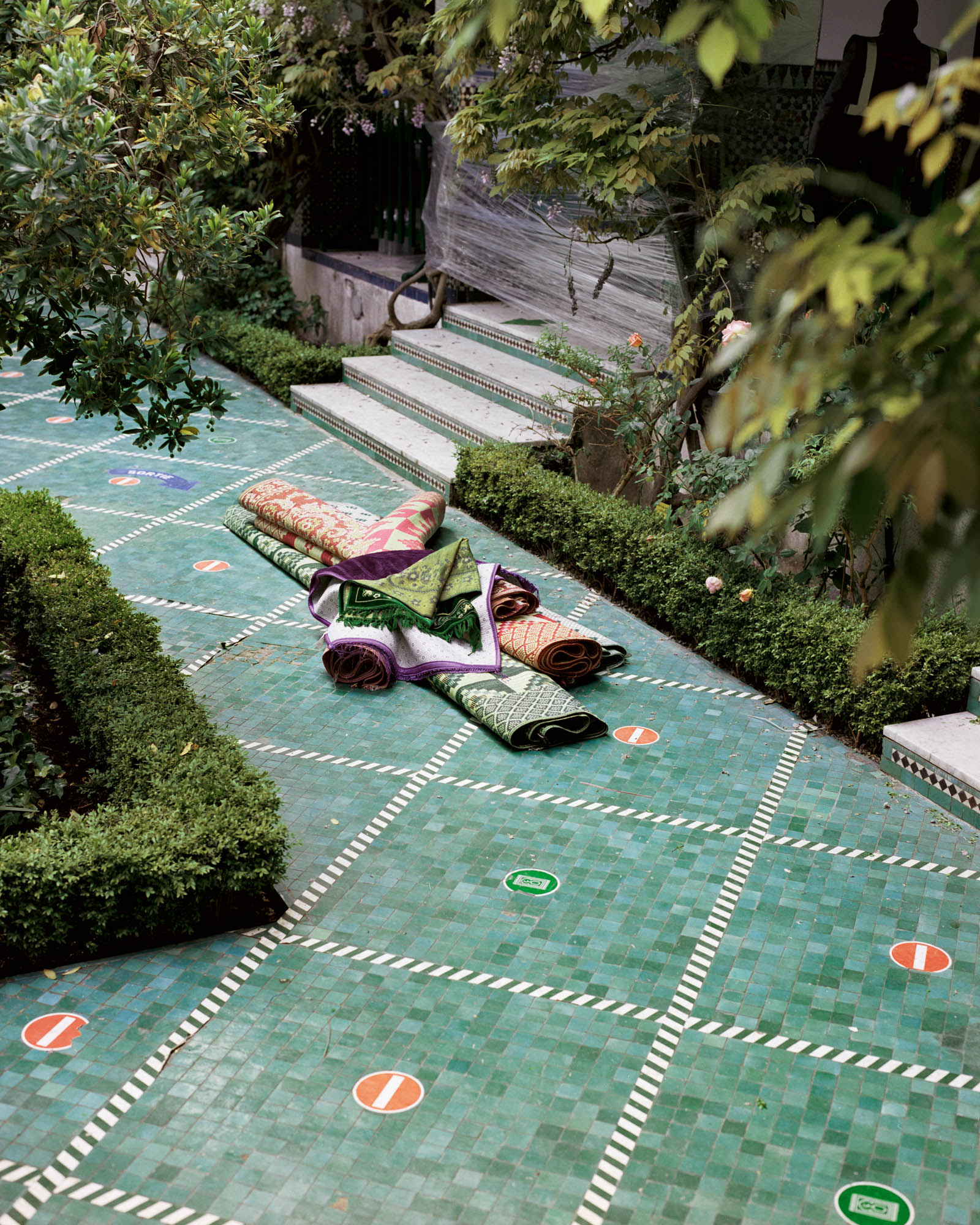 A photograph, titled Haram, of a pile of rolled up rugs on a tiled garden path. From Camille Farrah Lenain’s monograph Made Of Smokeless Fire