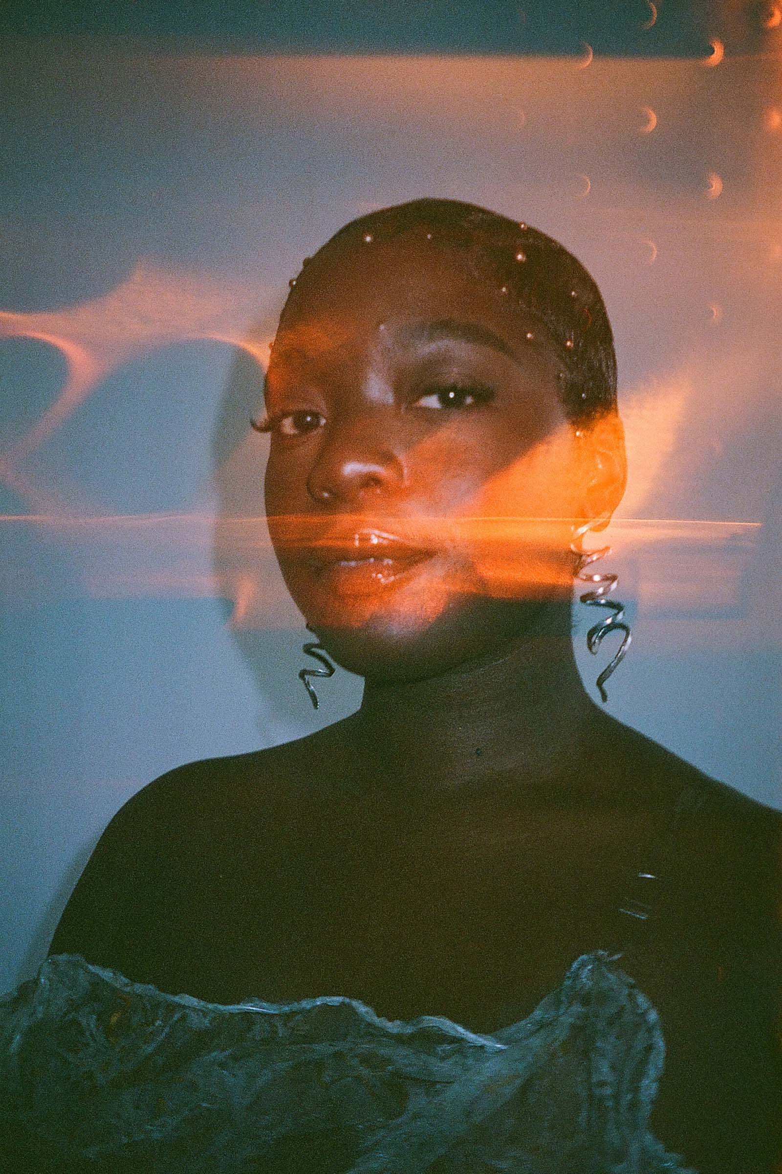 Photographer Ikram Abdulkadir's double exposure image overlays patches of orange light on a portrait of a woman wearing long curly metal earrings and a off the shoulder dress, looking directly at the camera