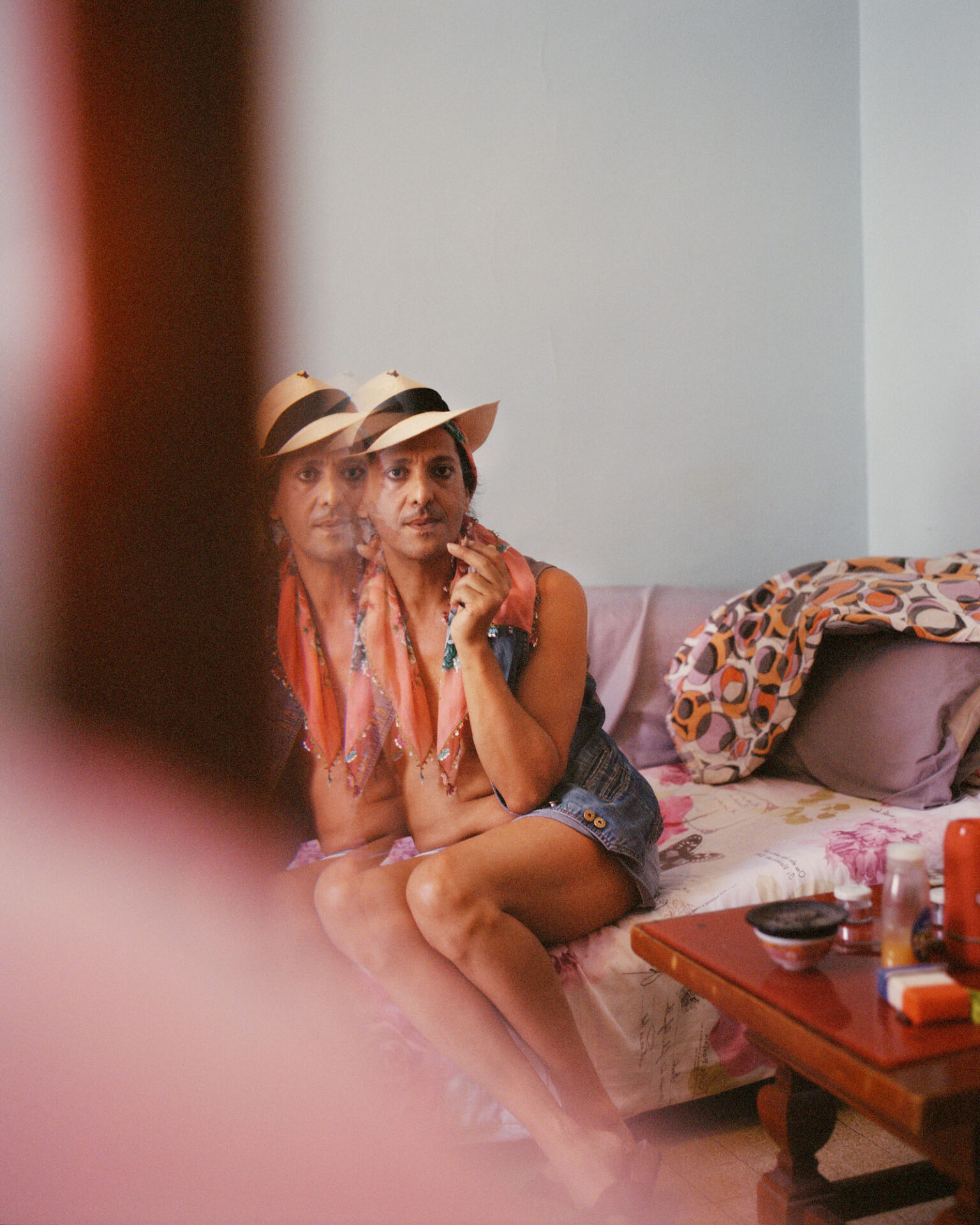 A double-exposed photograph, titled The mourning of the gays who have suffered, taken in Bouchta, Marseille in 2022, of a person wearing a hat and sitting on a sofa holding a cigarette, looking towards the camera. From Camille Farrah Lenain’s monograph Made Of Smokeless Fire