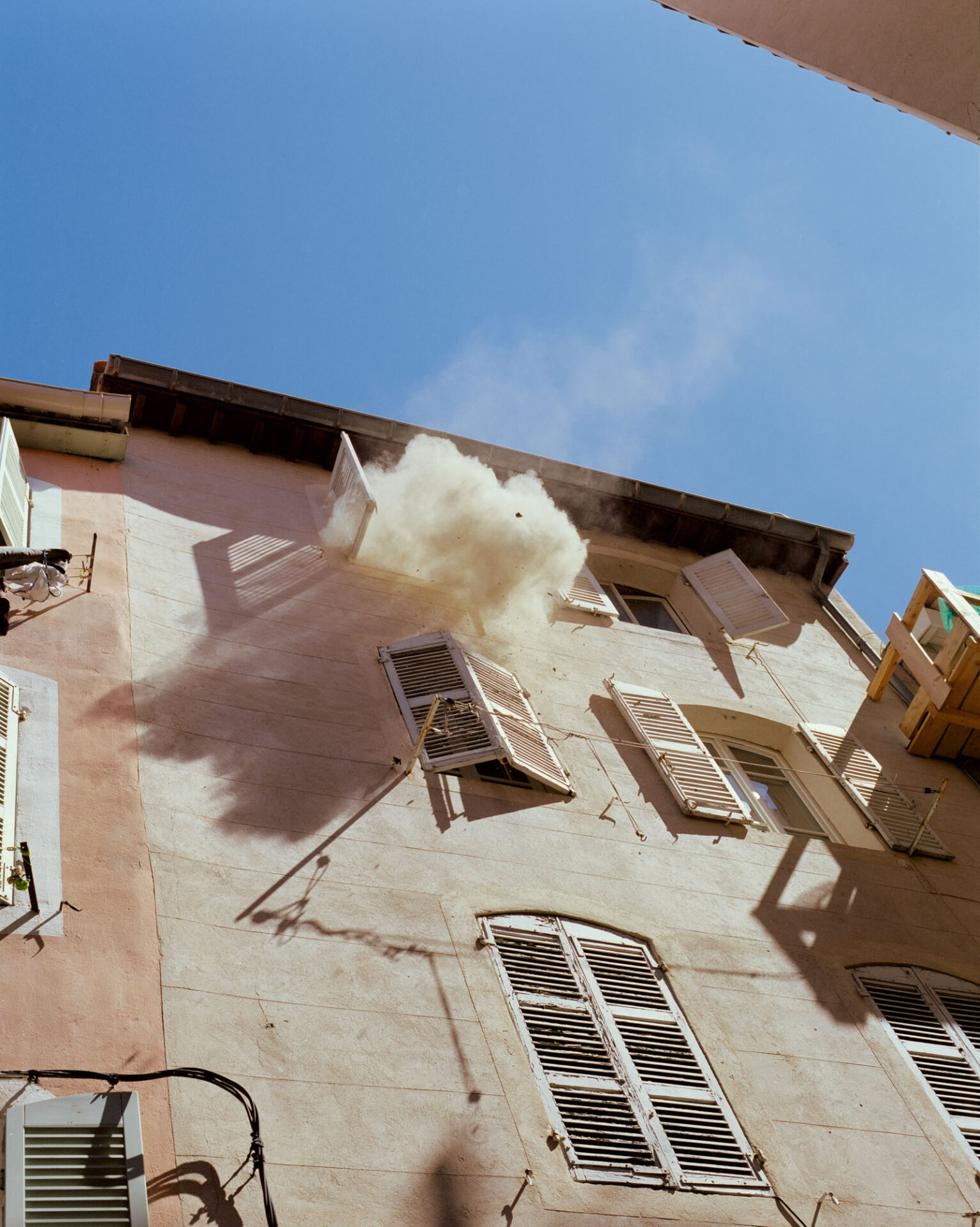 A photograph, titled Refuge, looking up at the side of a residential building as white smoke billows out of a top floor window. From Camille Farrah Lenain’s monograph Made Of Smokeless Fire