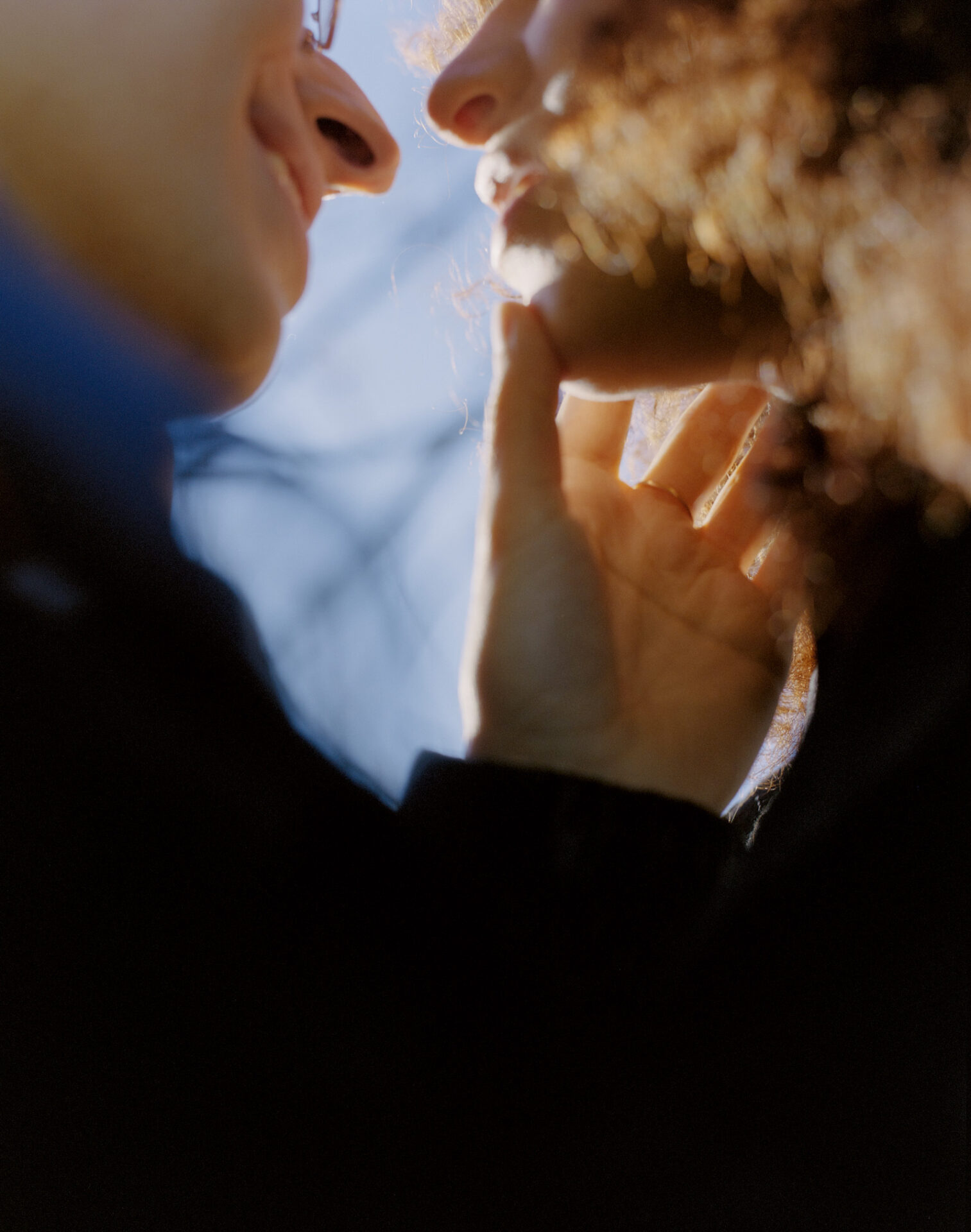 A close-up shot, titled N. and M. of two people apparently about to kiss, taken from below with their faces framed against the sun. The person on the left has placed their hand on the other's face, thumb on chin. From Camille Farrah Lenain’s monograph Made Of Smokeless Fire