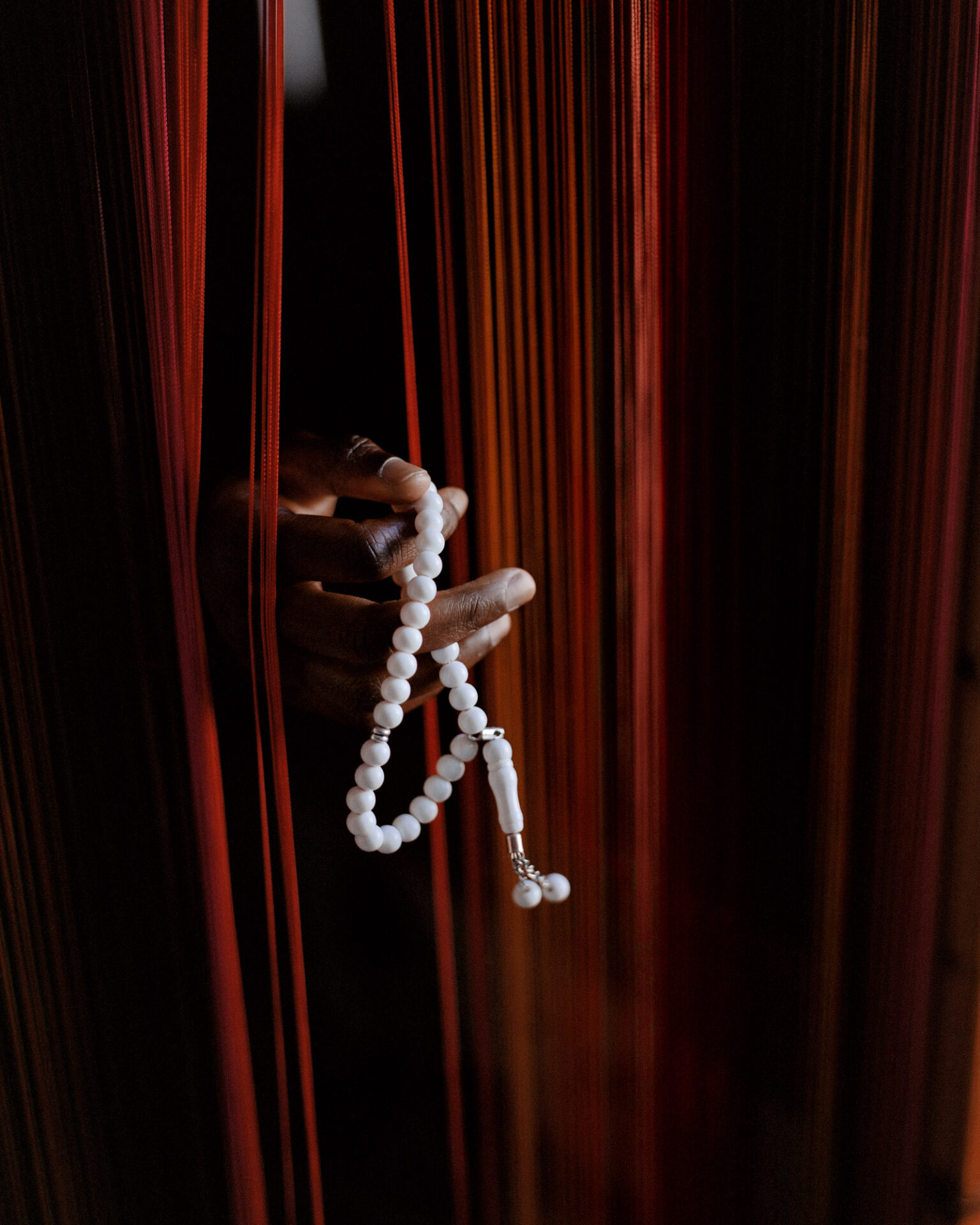 A portrait photograph, titled N., of a hand peeking out through a gap in red curtains, holding tasbih prayer beads. From Camille Farrah Lenain’s monograph Made Of Smokeless Fire