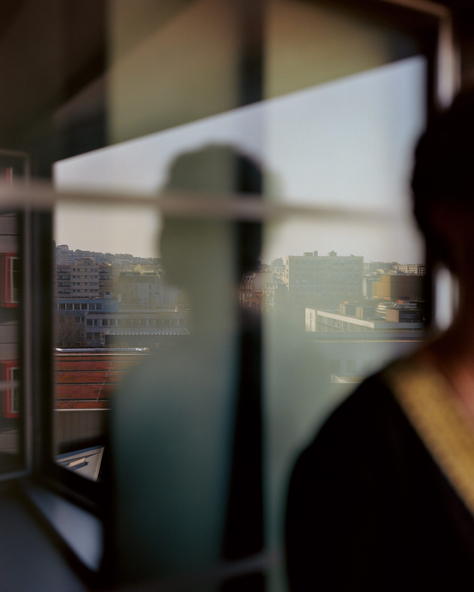 A photograph, titled Rizlaine Sayah, taken over the subject's blurred right in the foreground and showing them reflected from behind in a window, with an urban landscape visible through another window in the background. From Camille Farrah Lenain’s monograph Made Of Smokeless Fire