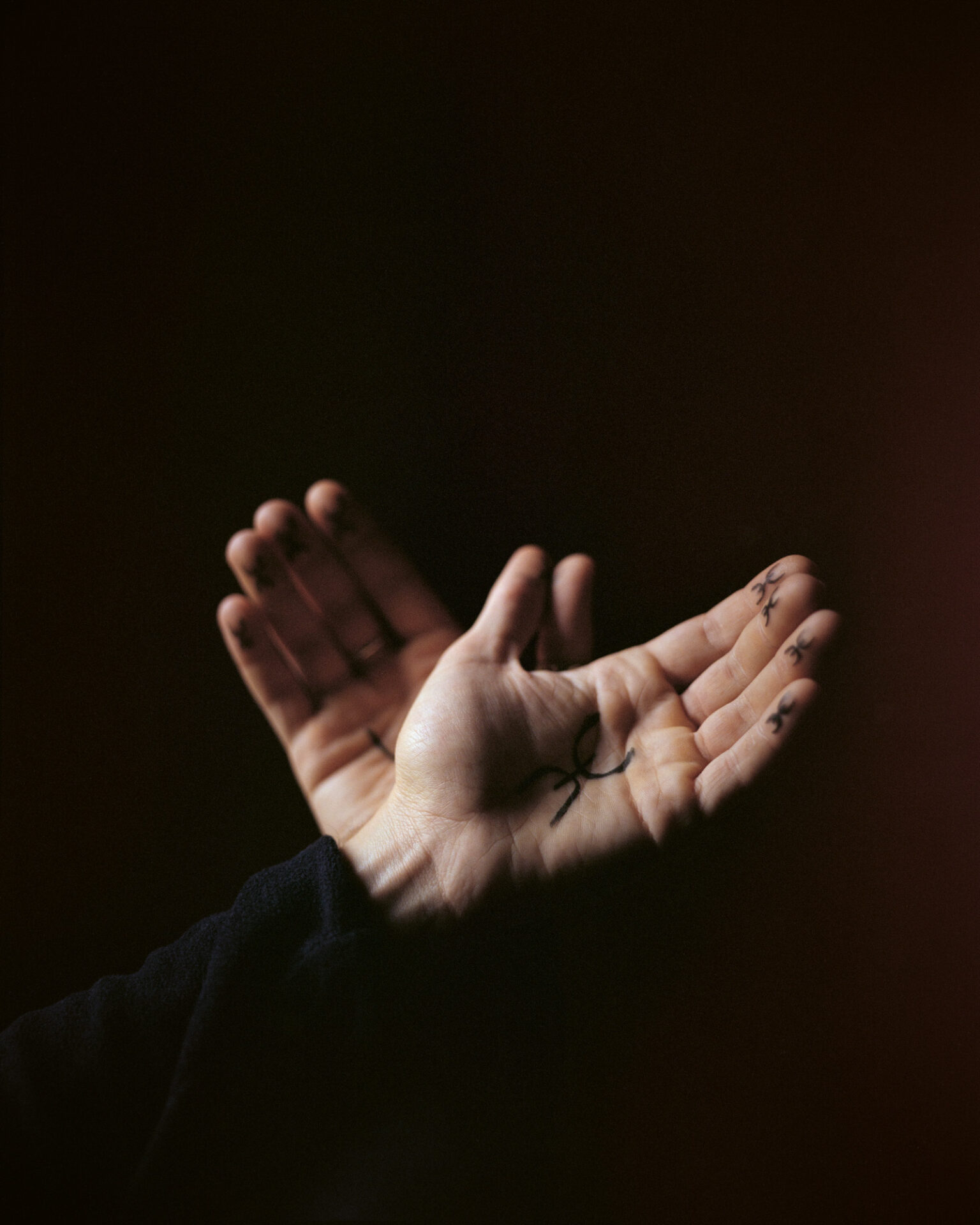 A photograph, titled ⵣ (Yaz), with two hands against a black background, each with a ⵣ symbol on each palm and finger. From Camille Farrah Lenain’s monograph Made Of Smokeless Fire