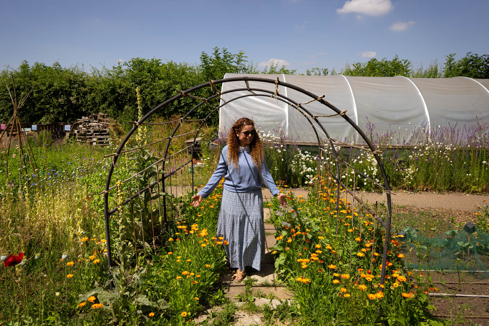 A photograph of a smiling woman, standing in bright sunshine with her arms outstretched and hands brushing flowers, in the Appletree Community Garden in Agbrigg, Wakefield