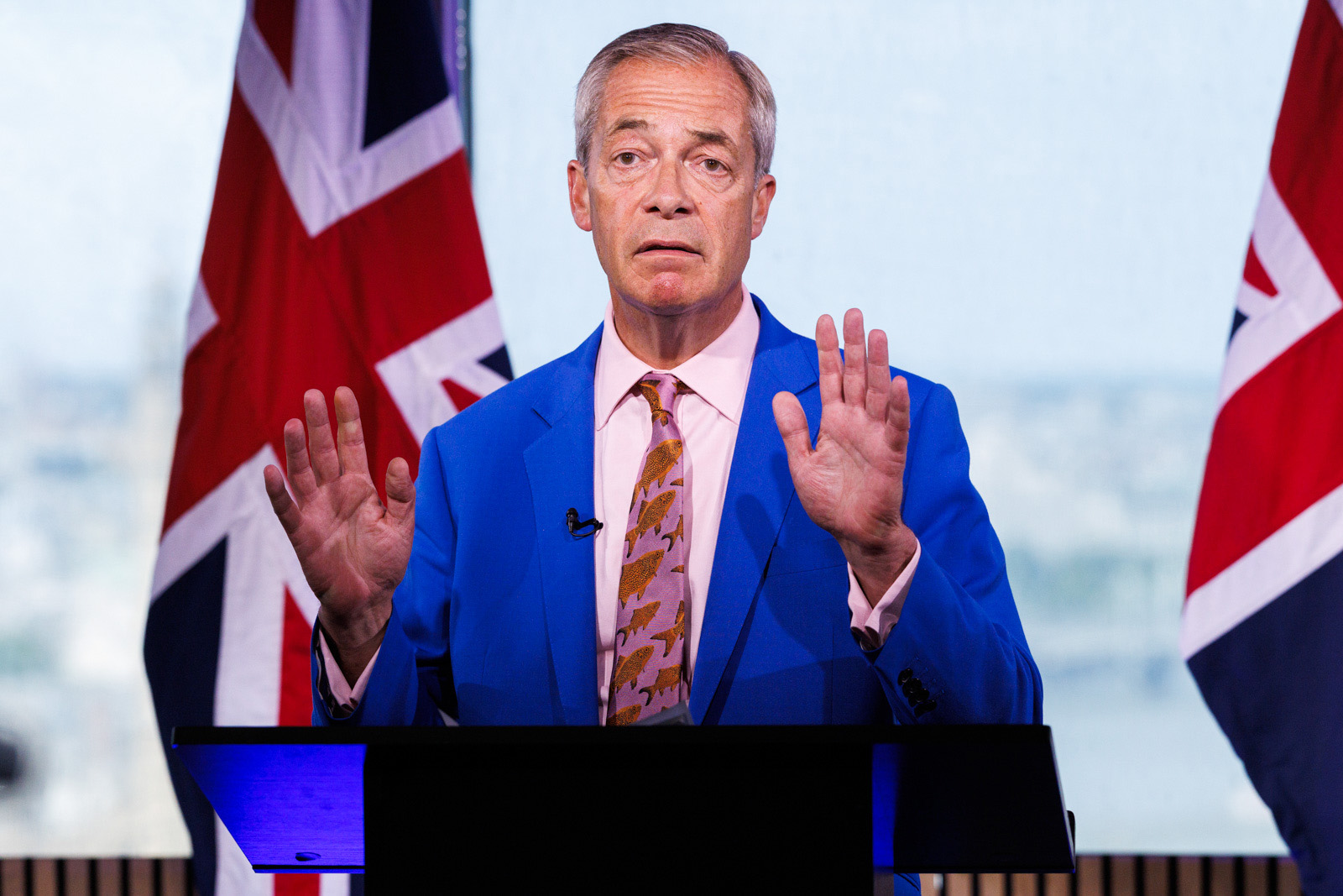 Nigel Farage flanked by union jack flags with a plaintive expression, raising both hands