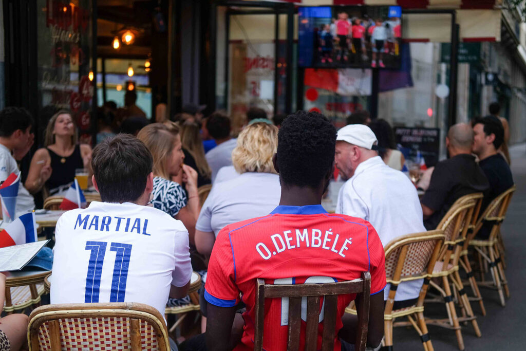 Two men, their backs to the camera, in a red O. Dembélé number 10 shirt and a white Martial number 11 shirt, seated watching a screen