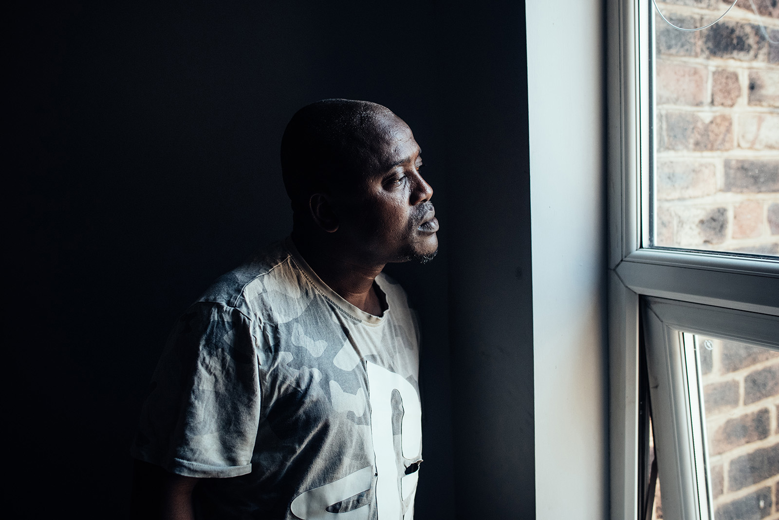 Alieu Barry photographed looking out of the window of his room in Stoke-on-Trent, July 2026