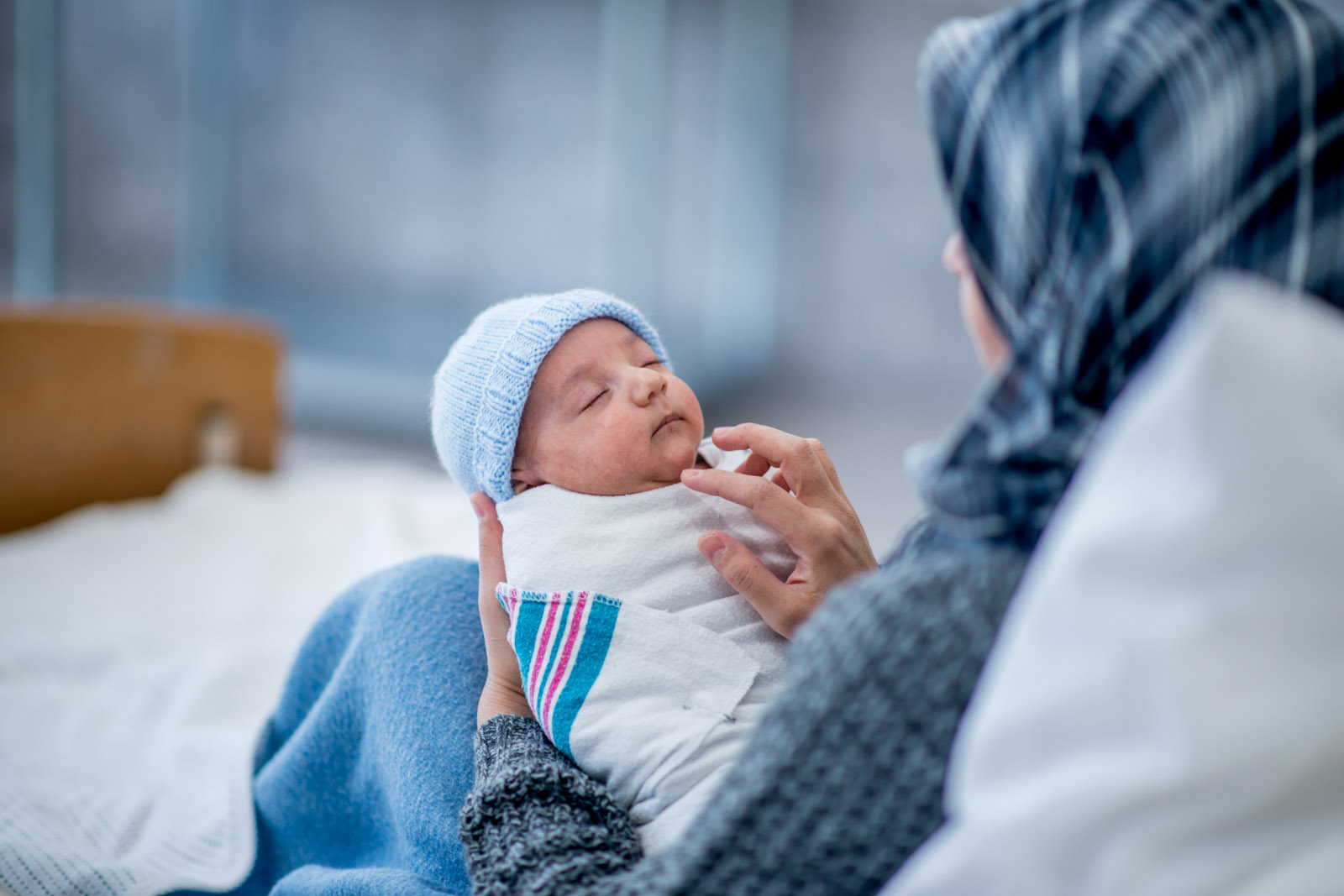 A stock photograph of a Muslim woman wearing a headscarf and sitting up in a hospital bed, holding her sleeping newborn son in her arms