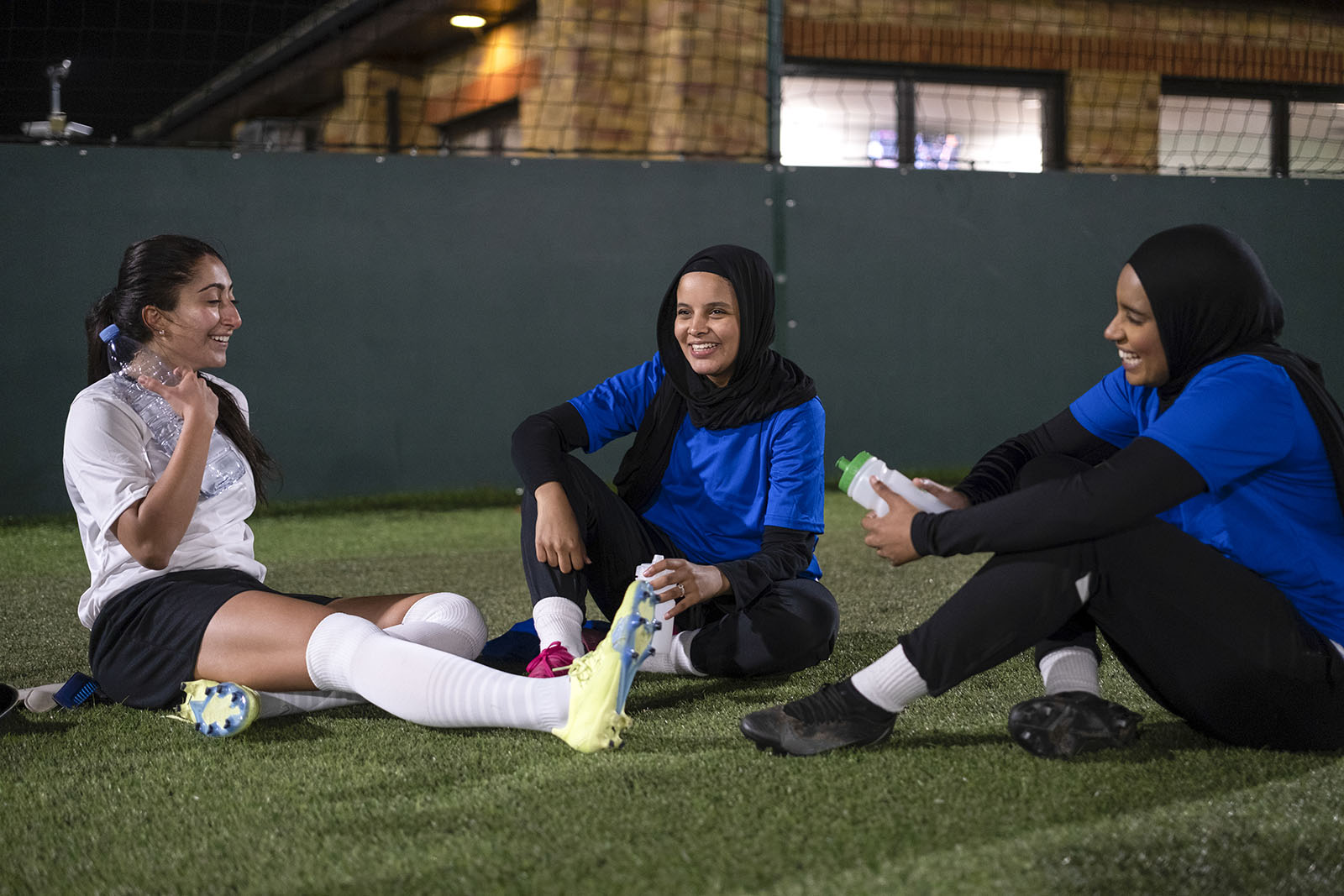 A stock image of three Muslim women football players, two wearing head scarves, sitting on the pitch smiling