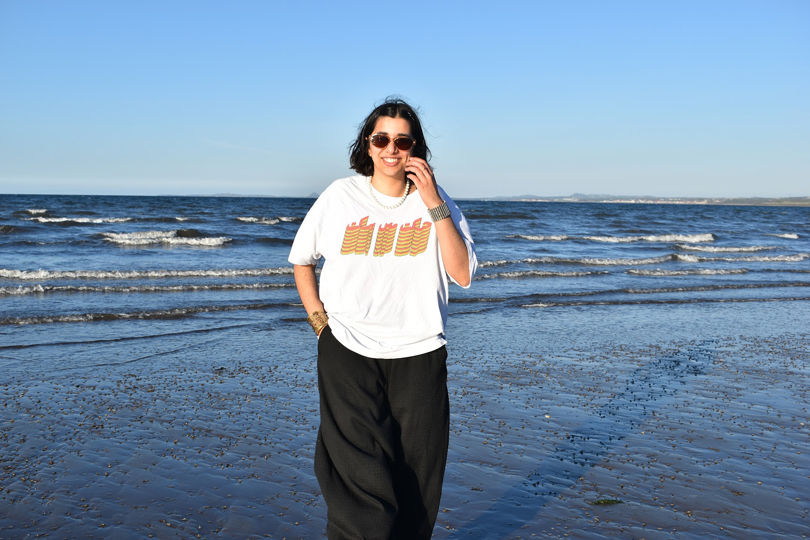 A photograph of Amina Aaliya Beg standing on a beach with the sea behind her and sun on her face, wearing sunglasses and a fundraiser T-shirt for Ajeeb Studio, which she set up with her husband James