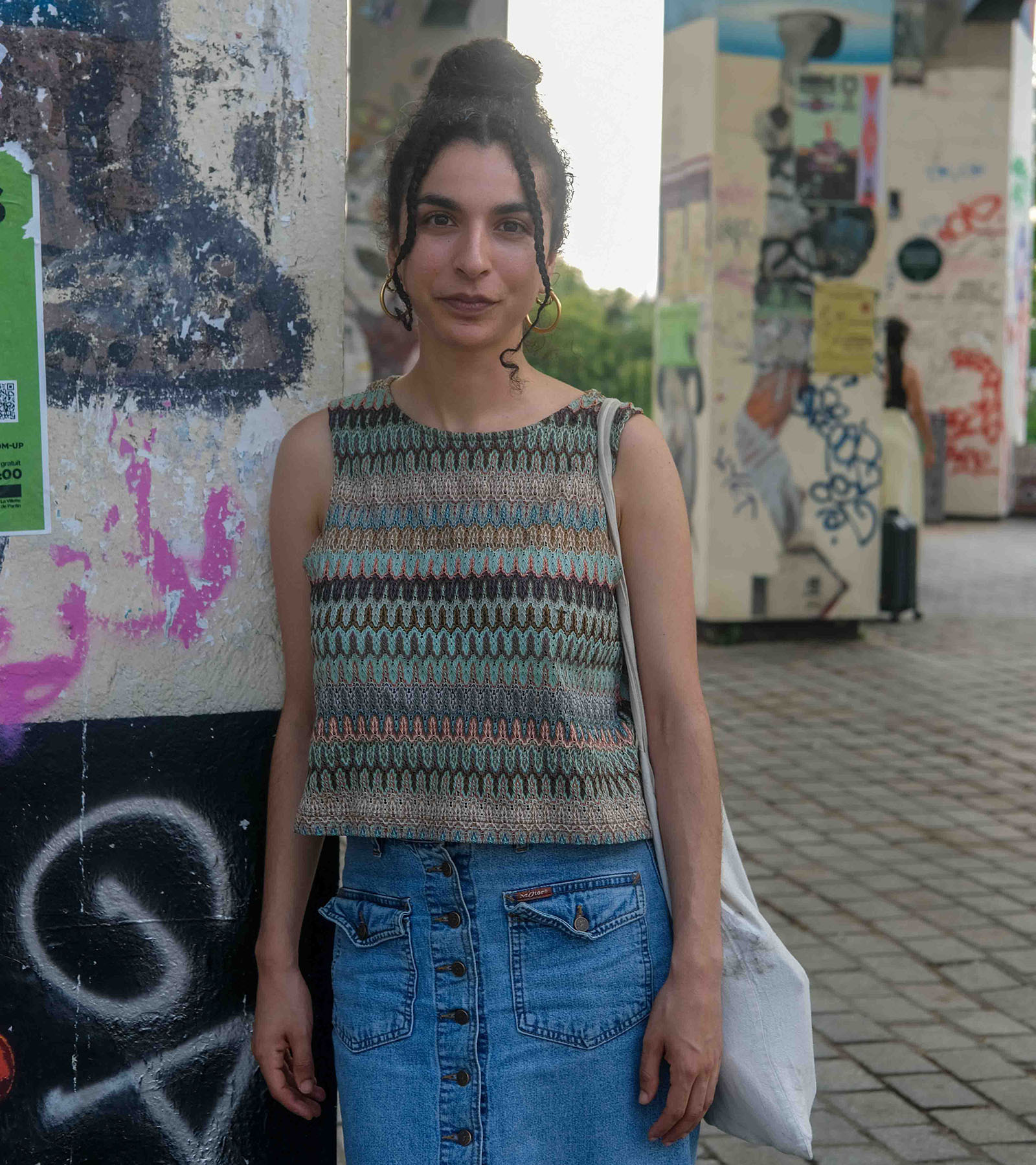 A woman with gold hoop earrings smiles for the camera. Behind her are concrete pillars covered with graffiti and the remains of posters