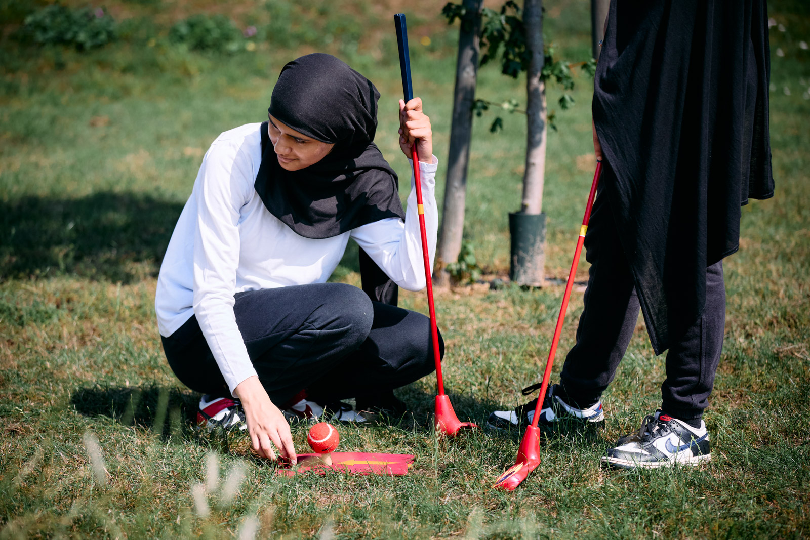 A photograph of Hareem Saeed in hijab kneeling on the grass holding a golf club and setting up a ball on a tee, during a Muslim Golf Association school event in Peterborough