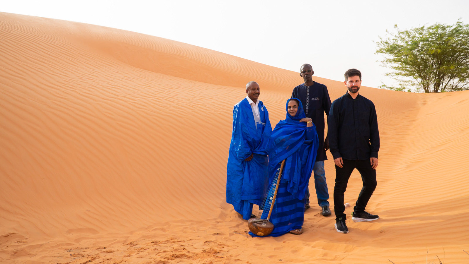 A publicity photo taken on a sand dune of Noura Mint Seymali with her band, husband and guitarist Jeich Ould Chighaly (left), bassist Ousemane Tour and drummer/producer Matthew Tinari (right)