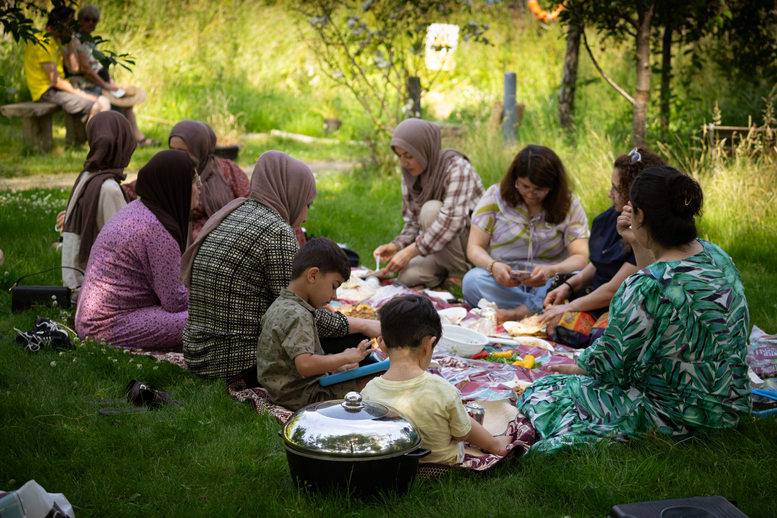 A photograph of women and children seated on a rug having a picnic in a shady patch of grass in the Appletree Community Garden.