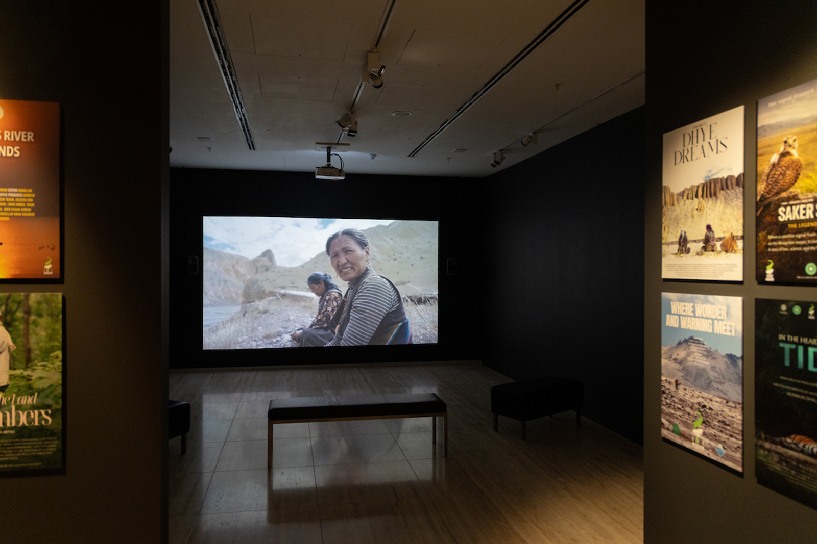 A photograph of the Voices from the Roof of the World exhibition at Aga Khan Centre Gallery, London, showing a darkened room with posters on the walls to the left and right and an image of a smiling woman, looking directly at the camera, projected on a large screen on the far wall