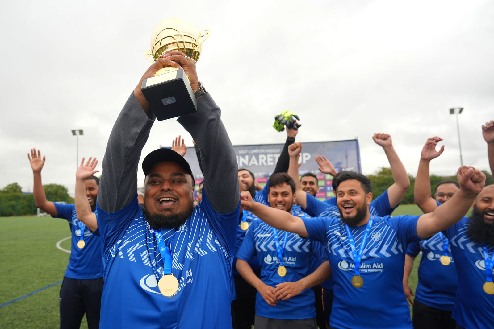 A photograph of the winning team wearing their medals and raising the trophy while celebrating victory in the the 2025 Minaret Cup, an annual football tournament that takes place at Mile End Stadium, London