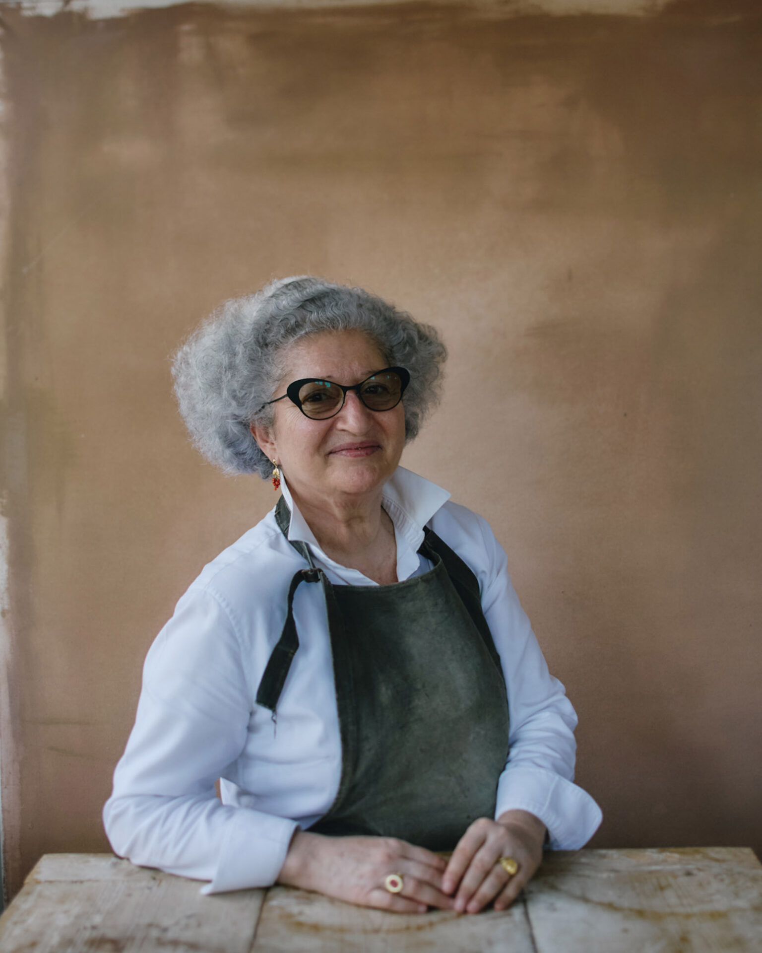 A portrait image of Anissa Helou, smiling and wearing an apron while seated at a table