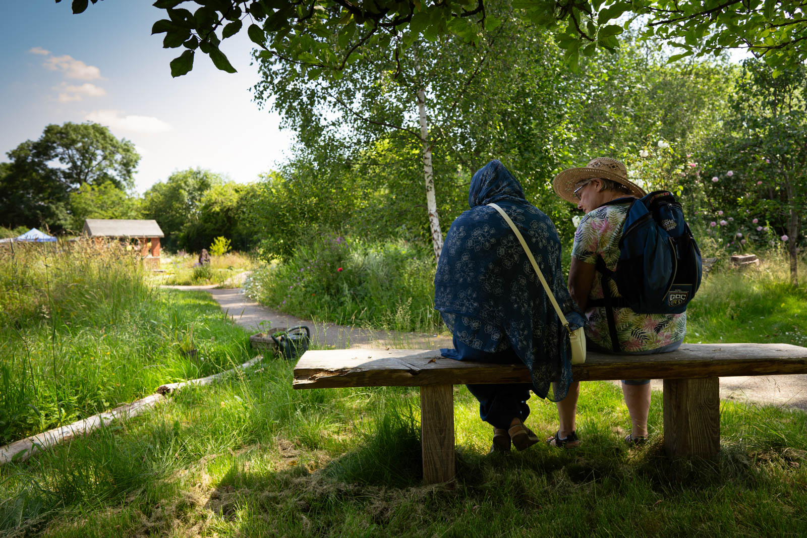A photograph of two women sitting on a bench under trees in a shady part of Appletree Community Garden