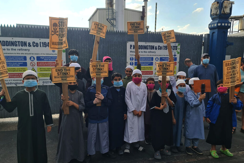 A photograph of a protest outside the P Waddington premises in Bradford, featuring a group of children and adults wearing face masks, with some holding 'End The Stink!' placards