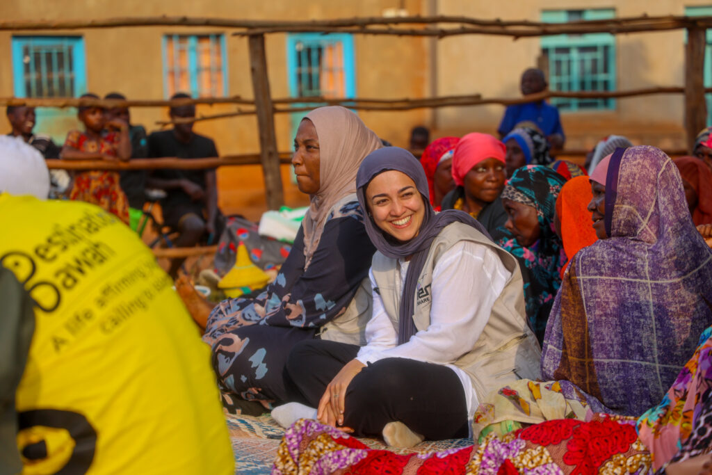A photograph of a smiling Mariam Amini, seated cross-legged in a group of local women, at a Destination Dawah Qur'an recital in Rwanda