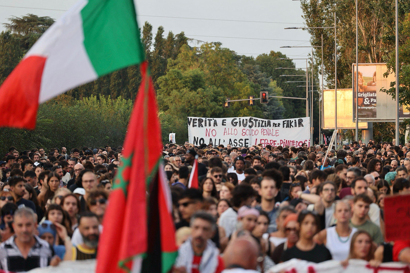 A photograph of a street in the Pilastro neighbourhood of Bologna on 26 July 2026, thronged with people marching in protest about the death of Abderrahim Fakir. Some are holding a large white banner with the 'Truth and justice for Fakir' daubed on it in black