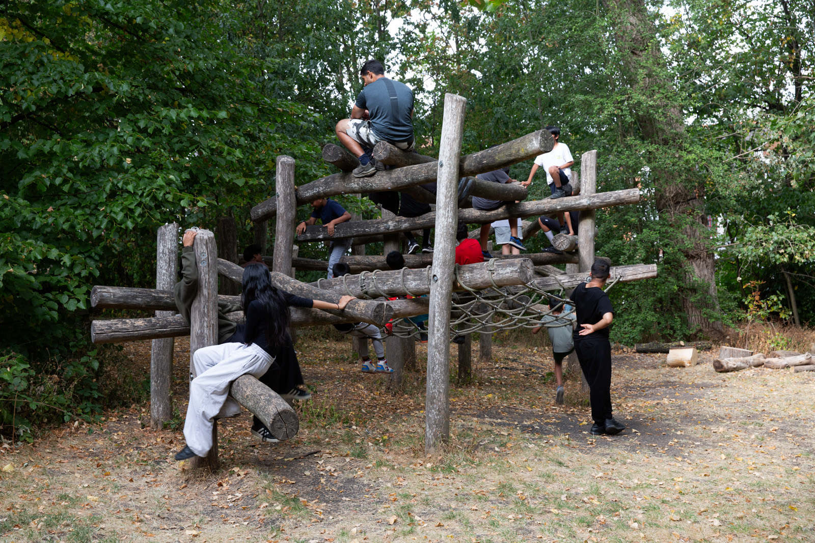 Children, their faces not visible, climb on a structure made of logs with trees in the background