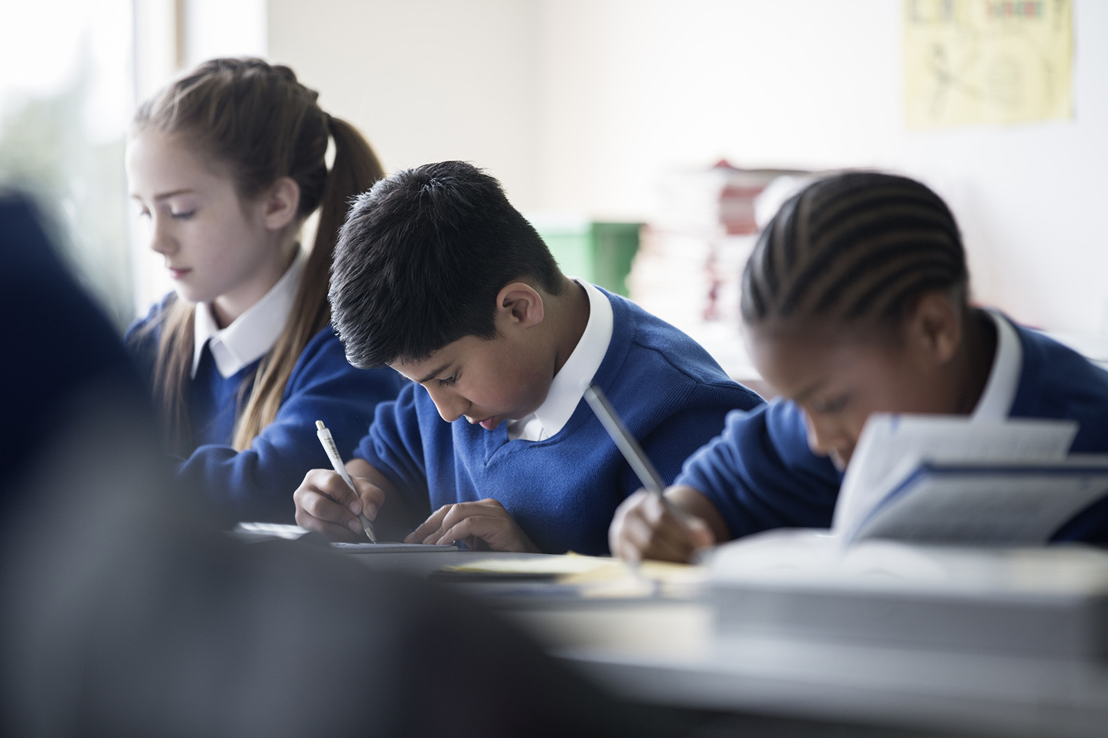 Stock image of three school children wearing matching blue jumpers sitting at their desks writing