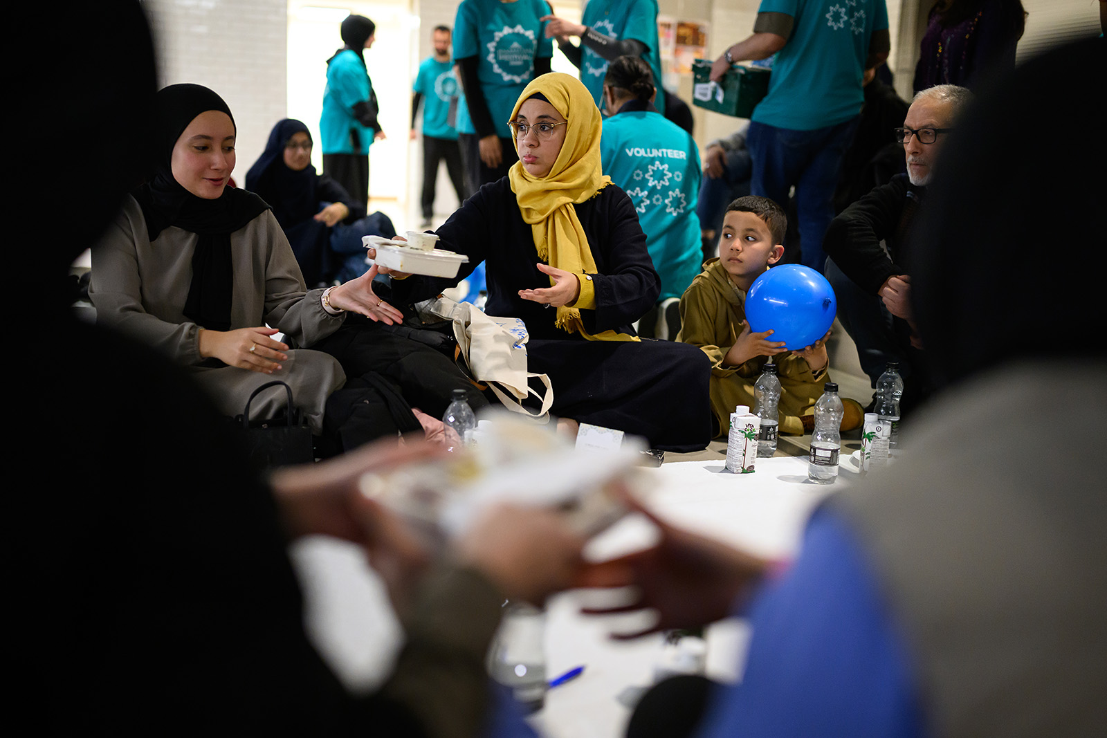 A woman in a yellow headscarf hands food to a woman sitting next to her. Other figures look on, surrounding a table with bottles of water