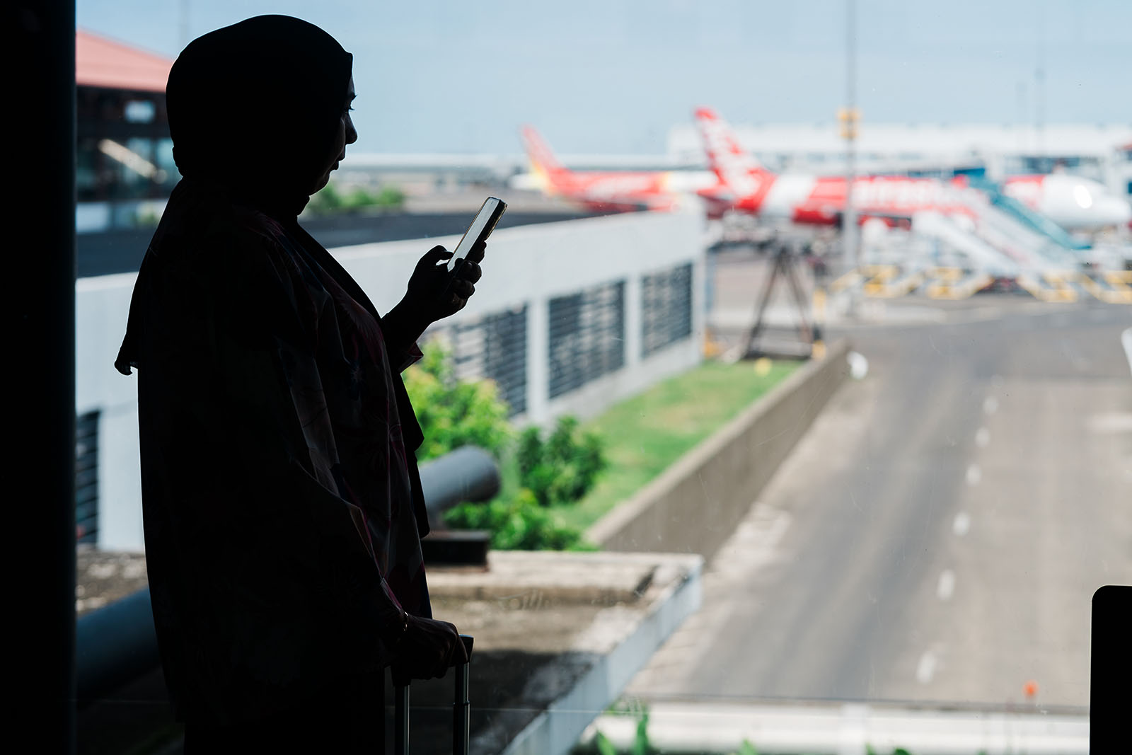 A stock image of a woman wearing a headscarf, shown in silhouette looking at a smartphone, in an airport terminal waiting to board her flight, with planes visible on the tarmac through a large window in the background.