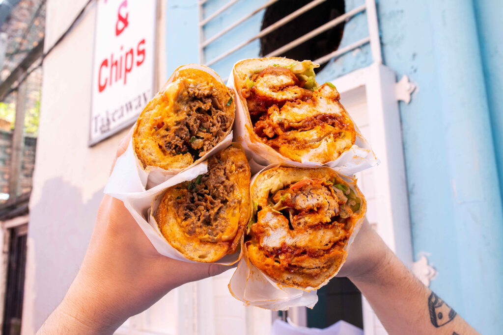 A close-up photograph of someone holding up halal sandwiches from Fat Pat's, Manchester, with fillings in the outlet's trademark eight-inch milk rolls, sliced in half and wrapped in paper