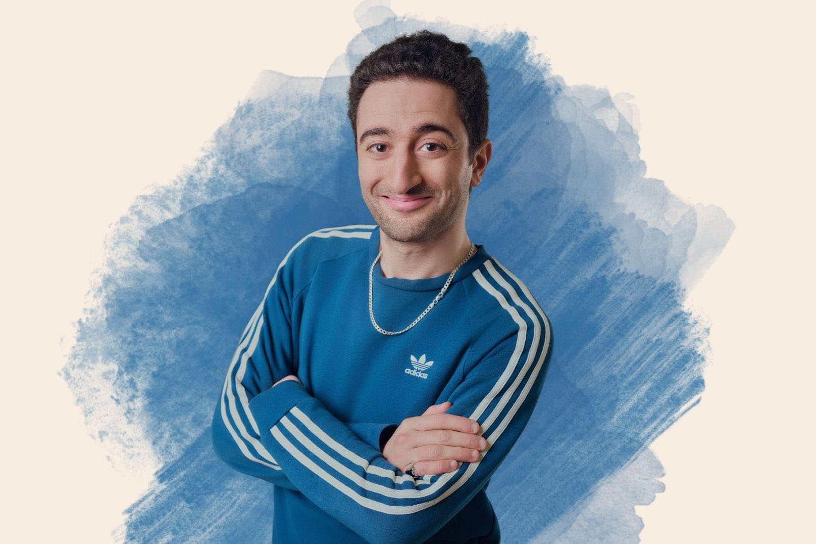 A portrait image of standup comedian Hasan Al-Habib, smiling with arms folded across a blue Adidas top