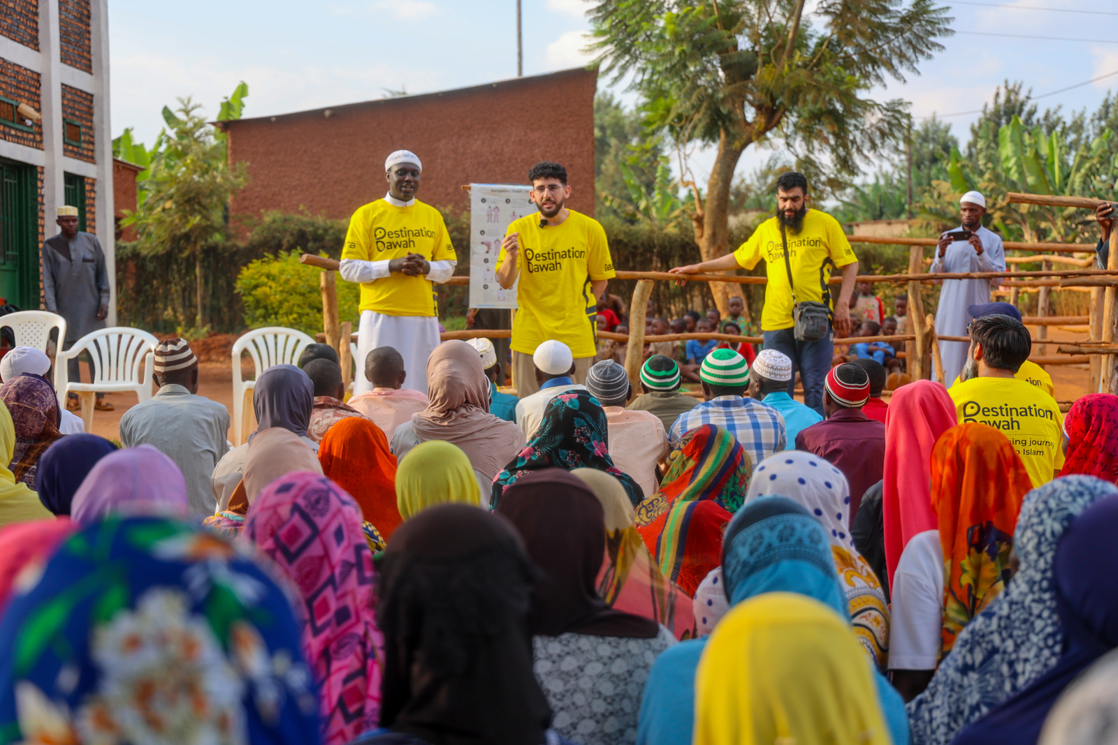 A photograph of a Destination Dawah Qur'an recital that Mariam Amini attended during her trip to Rwanda. The image shows three men, wearing bright yellow Destination Dawah T-shirts, addressing a crowd of men and women, including Amini, sitting on mats on the floor
