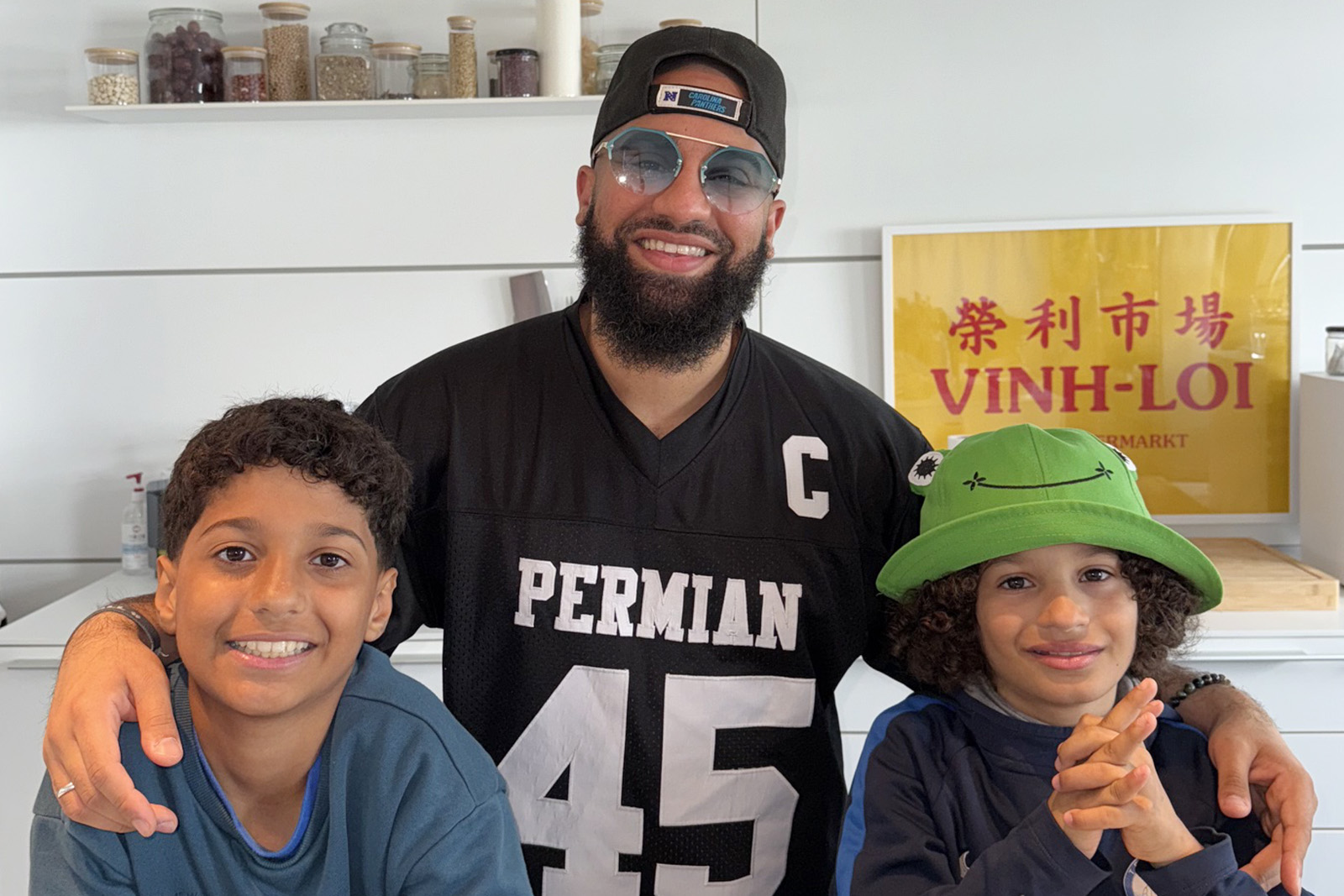 A photograph of a smiling Yassin Acosta Medina (AKA halal food influencer Yassin Halal), wearing a back to front baseball cap and with his arms around his two children, standing in what looks like a kitchen