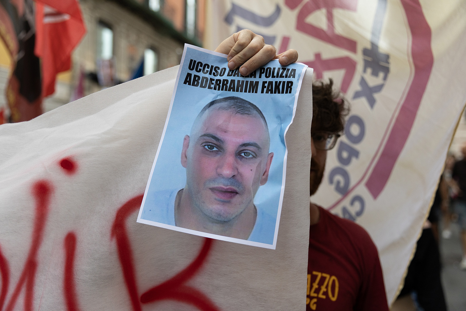 A photograph of a demonstrator holding an image of Abderrahim Fakir, with the words "killed by the police" printed on it, during a protest in Naples on 21 July 2026