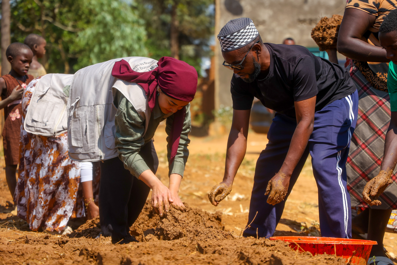 A photograph of Mariam Amini bending down with her hands on a pile of mud, while helping locals make bricks to build a home for a local widow in Kayovu village near Lake Rweru, Rwanda