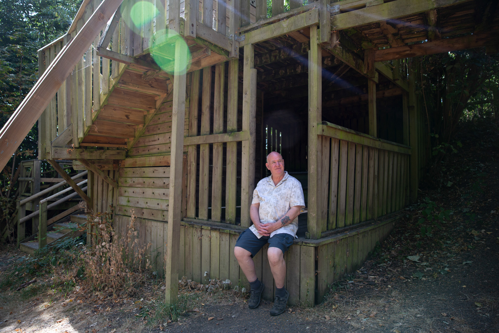 A man in a white patterned shirt sits on the step of a wooden play house