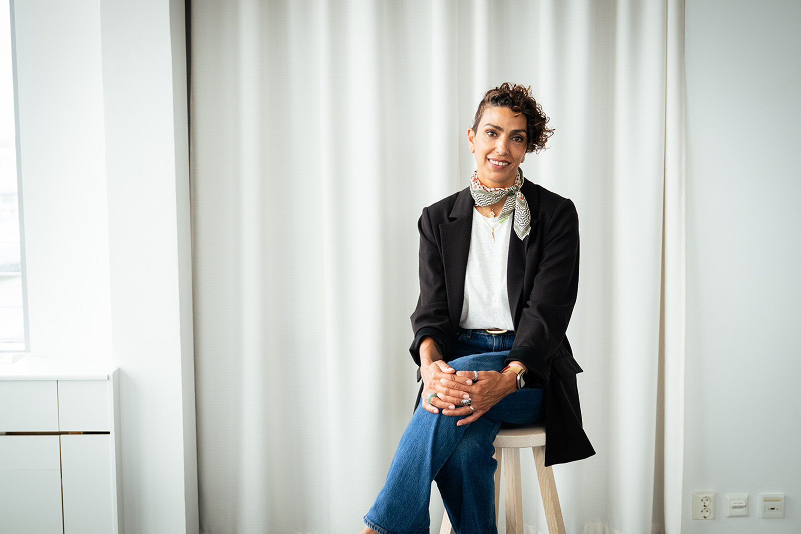 An interior portrait photograph taken in Stockholm in July 2026 of Linda Mutawi, seated on a stool with her hands clasped around her left knee and wearing a white top, black jacket and jeans