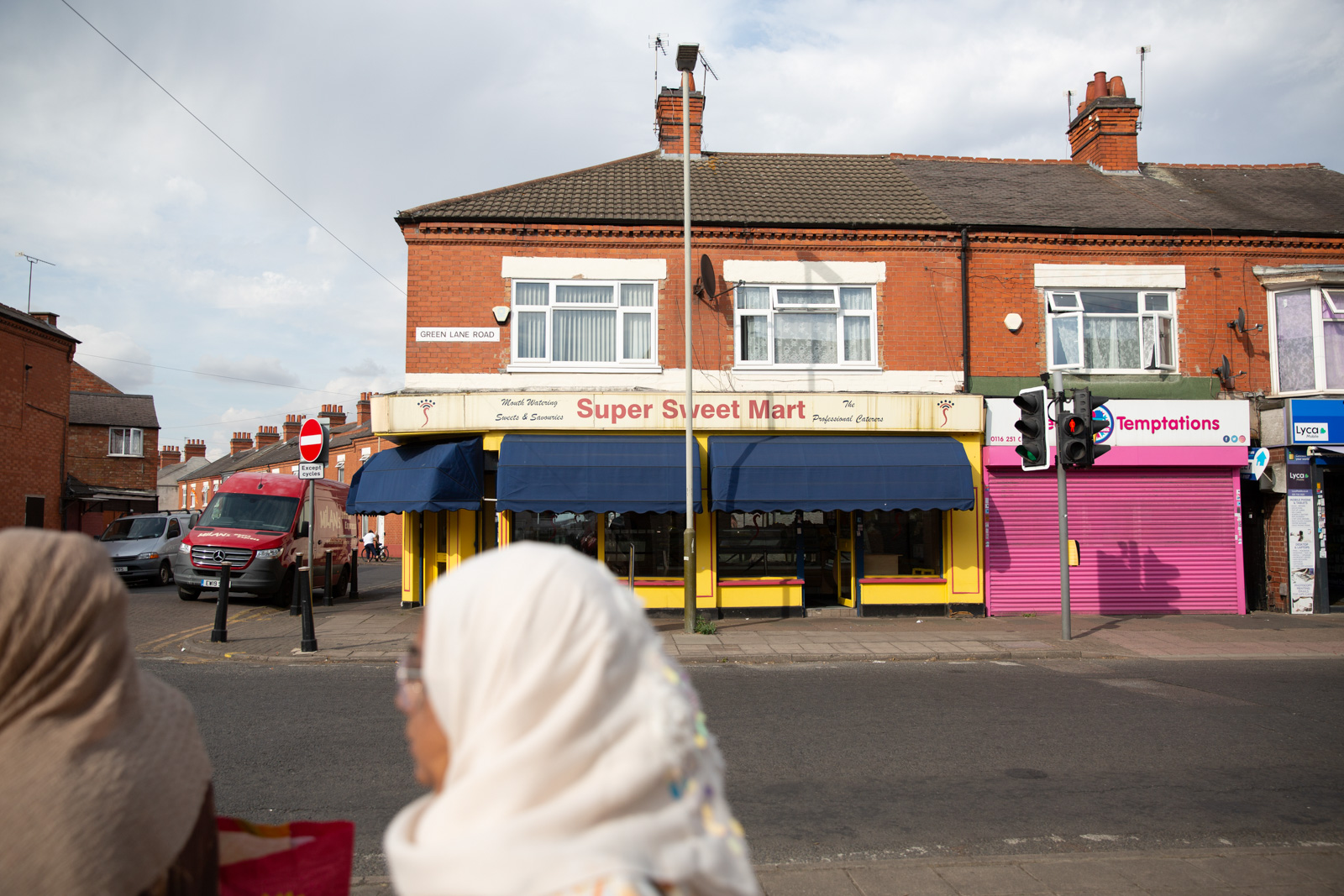 Shops, one reading 'Super Sweet Mart', along the bottom of a terrace. Across the street, nearer to the viewer, women in hijabs, their faces facing away from the camera, wait to cross the road