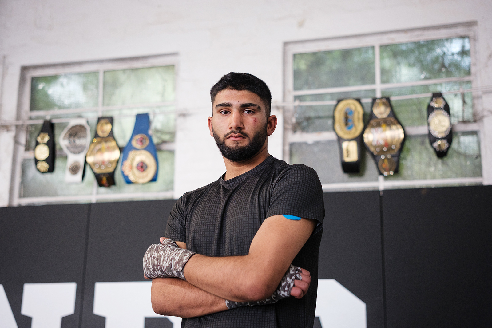 A portrait photograph of Muhammed Eesa, hands strapped and arms folded, standing in front of belts fighters from the gym have won at Legends MMA and Fitness Centre, Luton