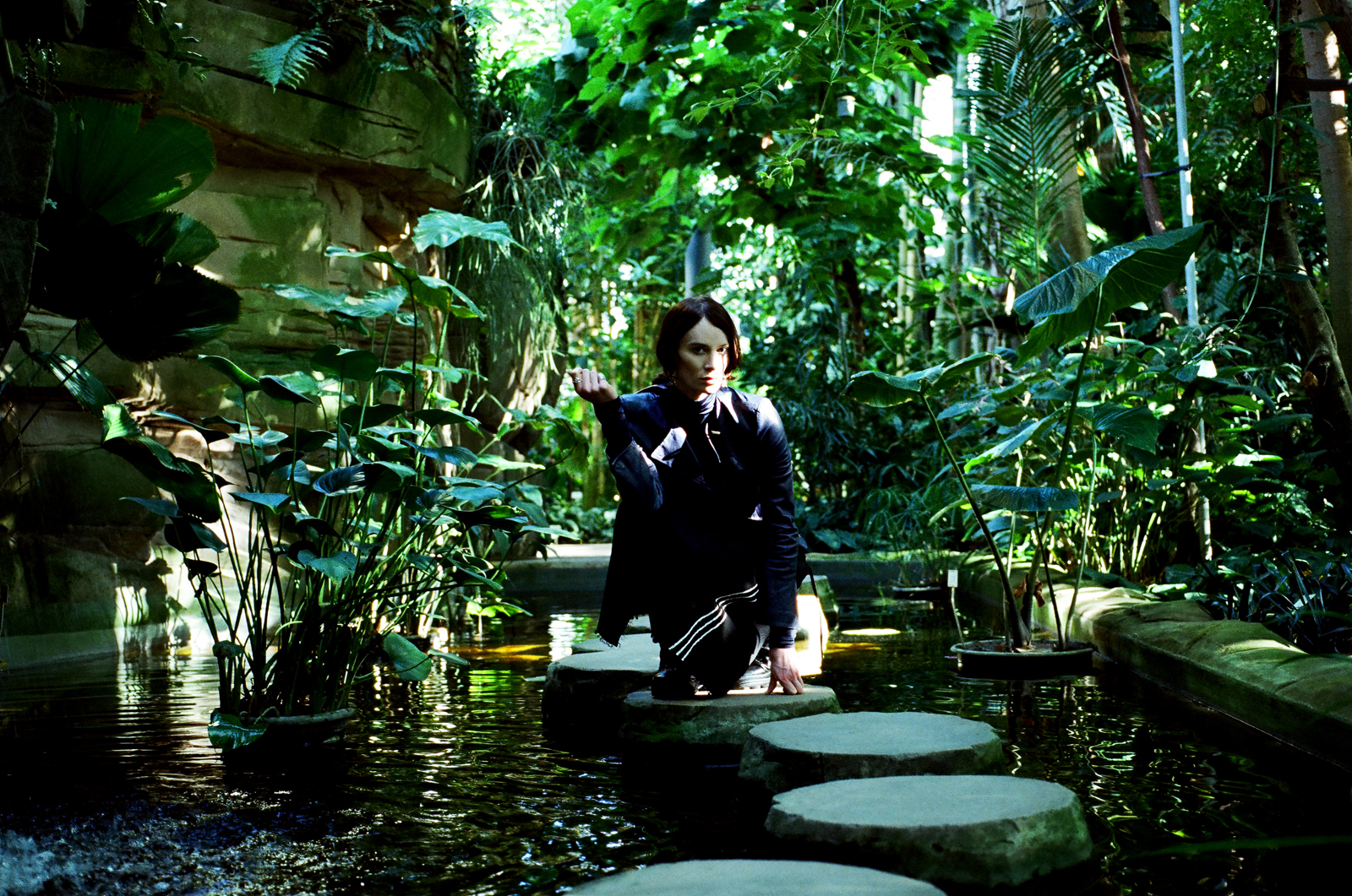 A promotional photograph of experimental musician Alif Hilal, who is kneeling on a stepping stone in a pond surrounded by foliage