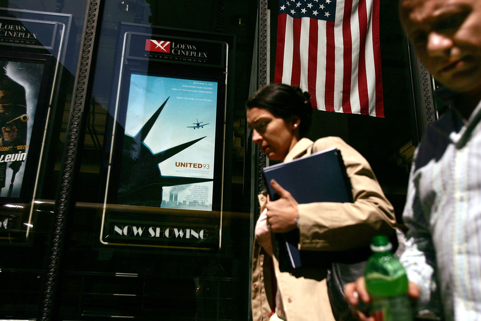 A photograph showing pedestrians — a man and a woman — walking past a poster for Paul Greengrass's 9/11 film United 93, beside a US flag, in New York, April 2006
