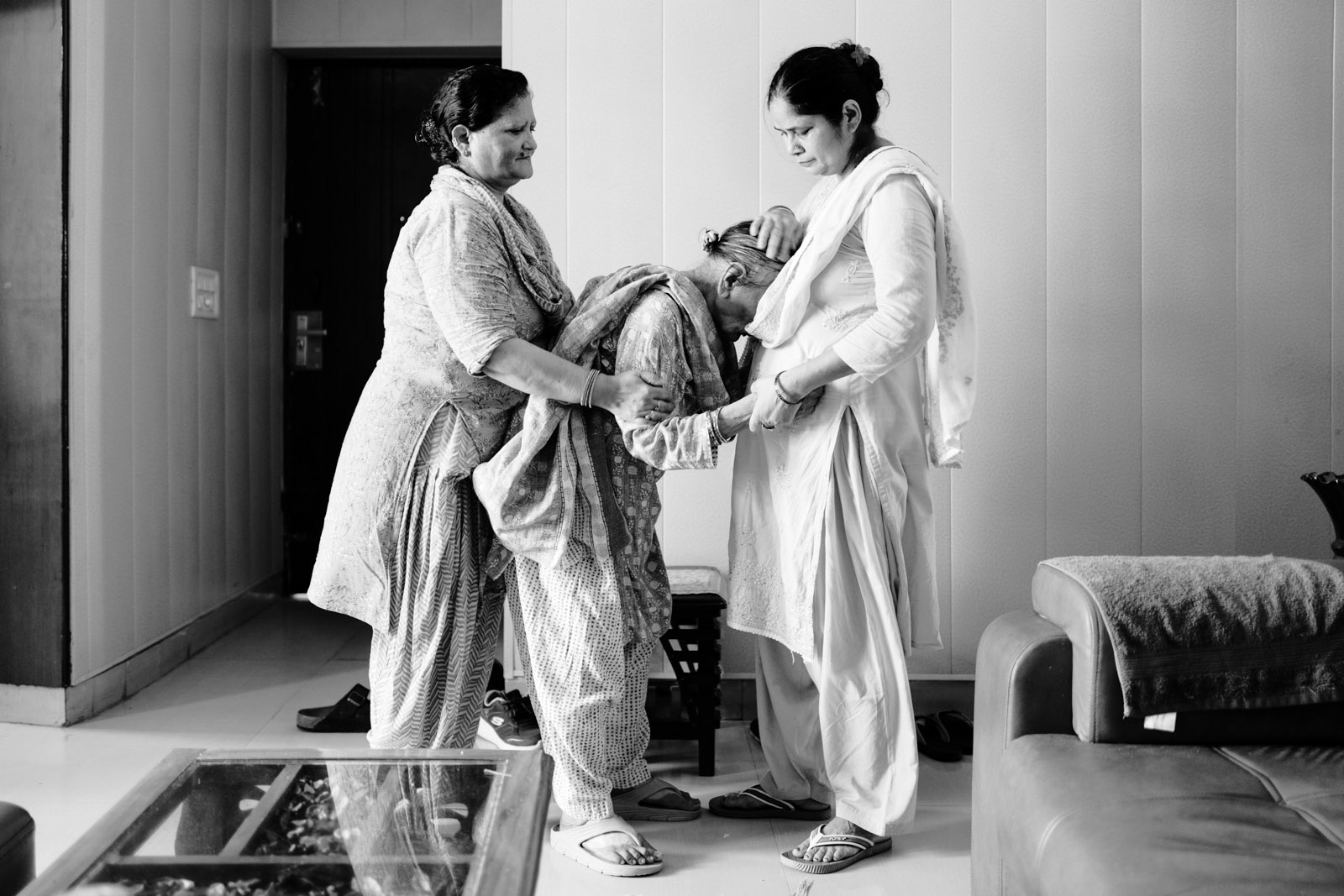 A black and white photograph from Aman Alam's series Ozymandias, documenting his grandmother's life with Alzheimer's, titled Naseem’s Morning Walk. In the image, Gudia and Laxmi, Naseem's two nurses, help her with her morning walk inside the house in Agra. She now needs help with the most basic of motor functions