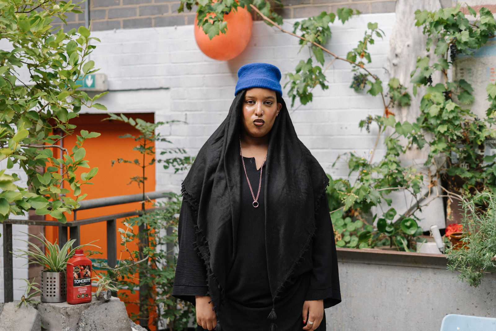 A photograph of artist Asmaa Jama in her studio in the Spike Island neighbourhood of Bristol. Jama is standing in front of a white-painted brick wall with an orange door to the left, surrounded by plants and wearing a blue knitted beanie hat, black shawl and a long-sleeved black dress