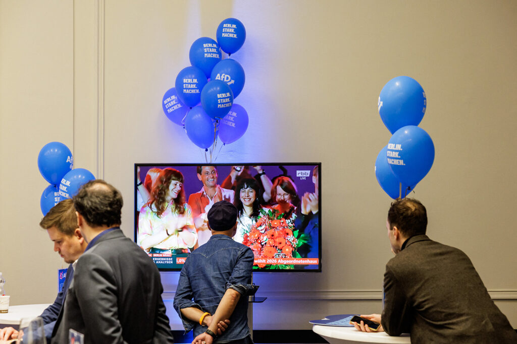 A photograph of an AfD Berlin state election night event, with a couple of the far-right party's supporters watching a TV screen showing Left Party leader Elif Eralp celebrating victory on 20 September 2026