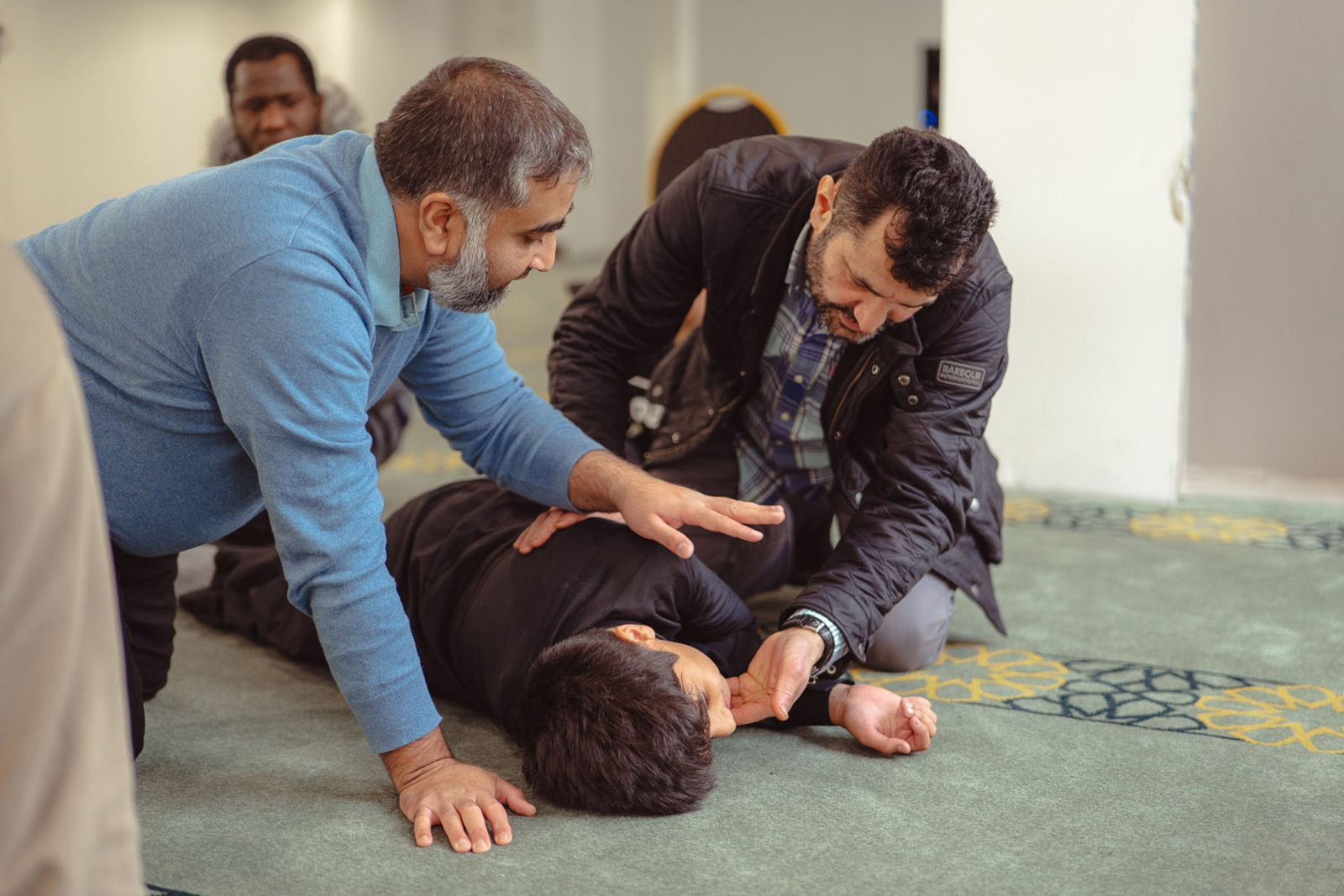 Two men kneel beside a man lying in the recovery position, one gesturing as if instructing the other