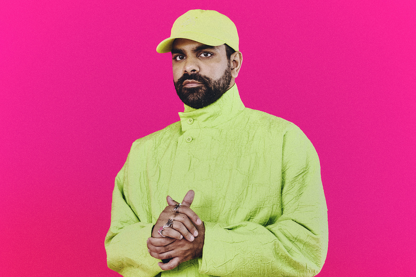 A portrait photograph of DJ and producer Ahadadream, wearing a bright lime green jacket and baseball cap against a pink background