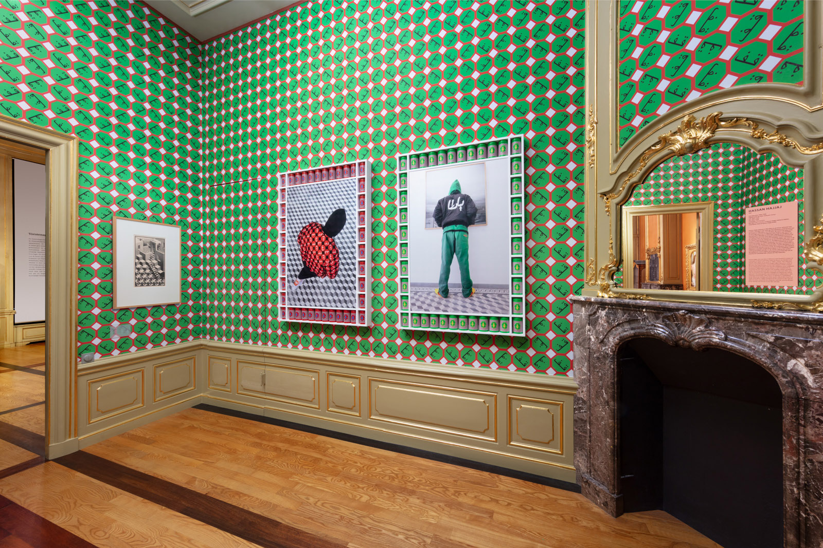 A photograph of the Escher and Islamic Art exhibition in the Hague, featuring a room with a large, ornate fireplace and wallpaper covered with bright green circles with a repeated Arabic phrase inside. An Escher picture is hanging on the wall alongside two images of people wearing hoodies with their backs to the camera, standing on floor covered with an Escher pattern