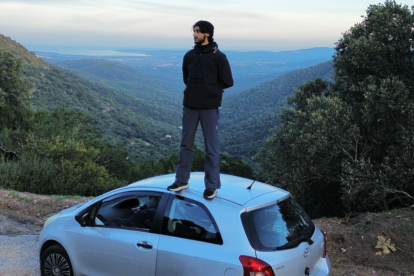 A photograph of Haris Tagari standing on top of his Toyota Yaris with a hilly, heavily forested landscape behind him and the coast in the distance, in Normandy, France on an earlier trip